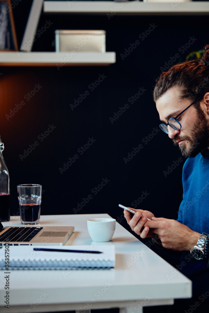 Side view of serious hipster sitting in his office and using smart phone. Side view of serious hipster sitting in his office and using smart phone.