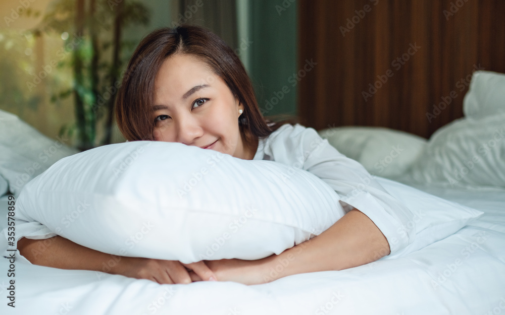 A beautiful asian woman holding a pillow while lying down on a white cozy bed at home A beautiful asian woman holding a pillow while lying down on a white cozy bed at home