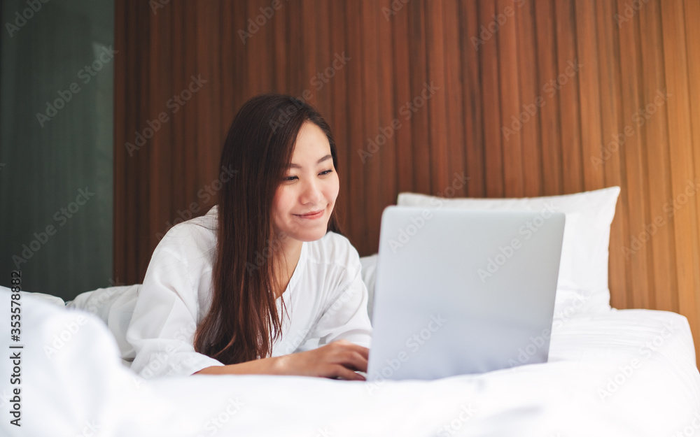 A beautiful asian woman using and working on laptop computer while lying down on a white cozy bed at home A beautiful asian woman using and working on laptop computer while lying down on a white cozy bed at home
