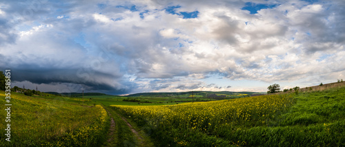 Spring yellow flowering rapeseed fields  ground road  dramatic cloudy sky with circular thunderstorm cloud and green rural hills. Natural seasonal  farming  climate  countryside beauty concept scene.