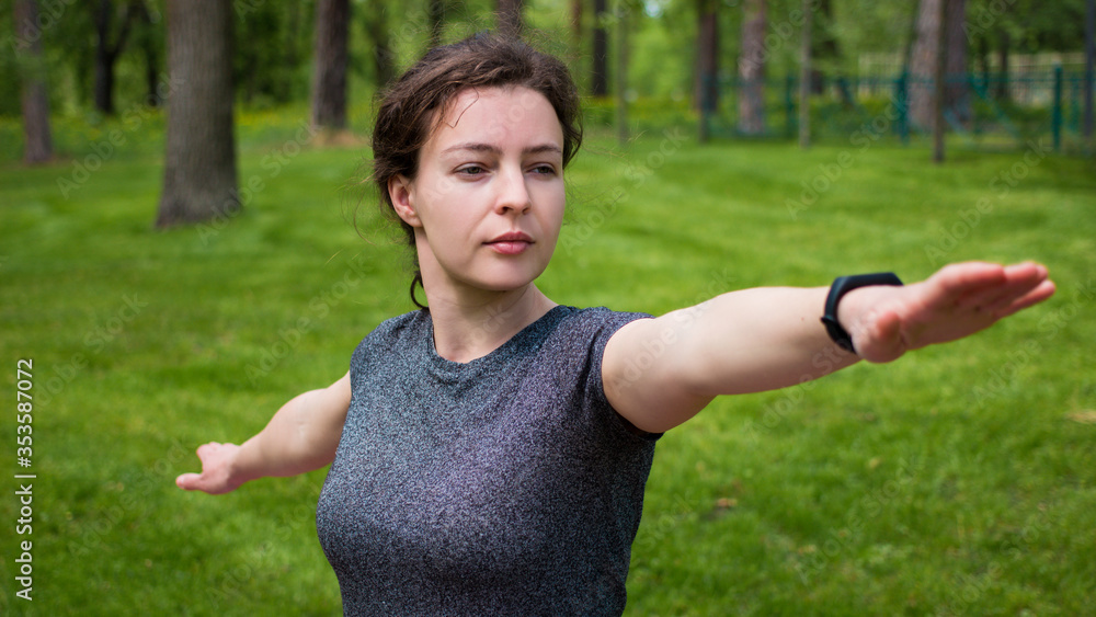 Young caucasian woman practicing yoga standing standing in Warrior two exercise,Virabhadrasana pose in green grass city park. Athletic girl in grey sport t-shirt training stretching work out Young caucasian woman practicing yoga standing standing in Warrior two exercise,Virabhadrasana pose in green grass city park. Athletic girl in grey sport t-shirt training stretching work out