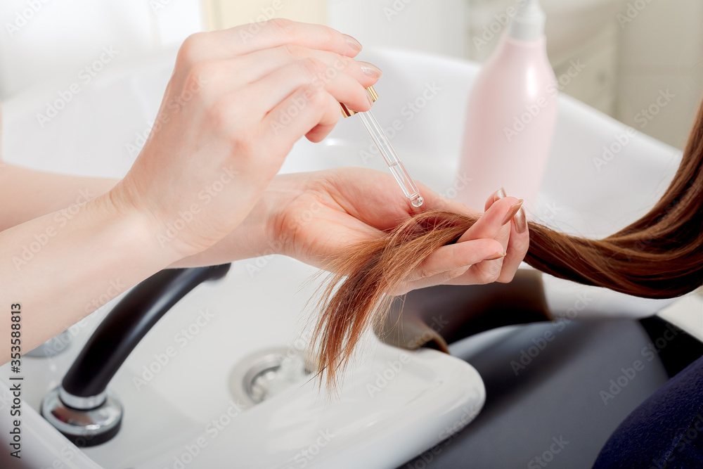Female hands applying oil on long womans brown hair. Hair care cosmetics, bath beauty spa products. Female hair serum for treatment hair tips against beauty salon interior, bathroom Female hands applying oil on long womans brown hair. Hair care cosmetics, bath beauty spa products. Female hair serum for treatment hair tips against beauty salon interior, bathroom