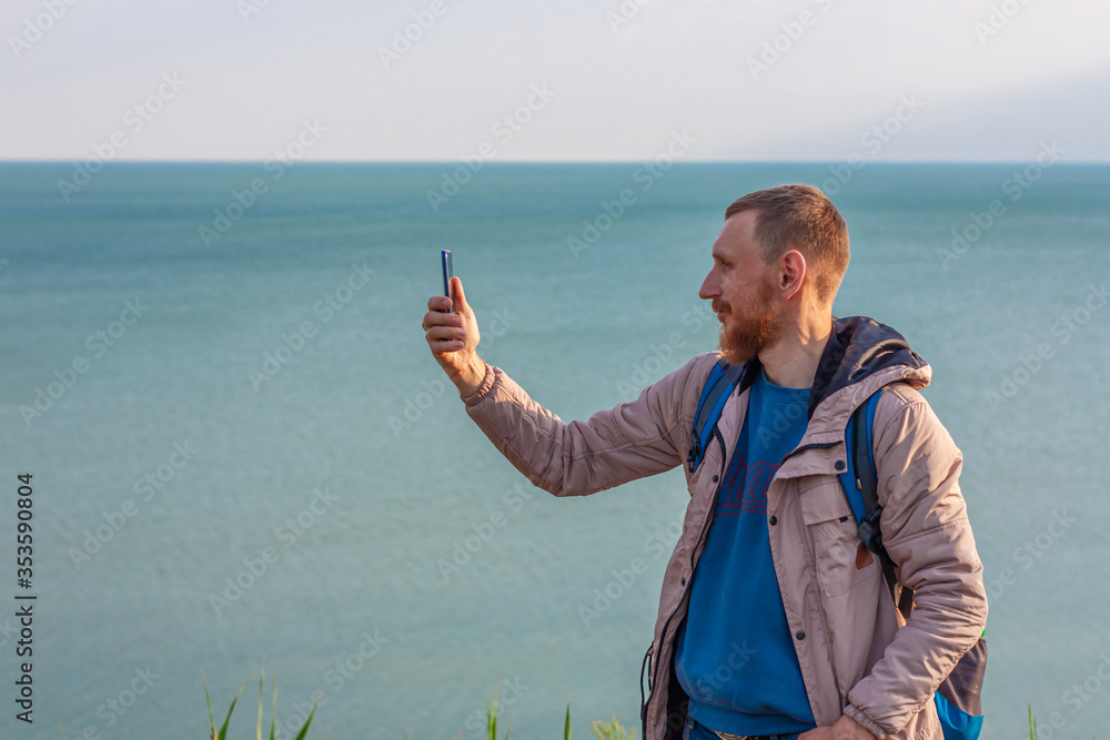 Bearded man in light raincoat on a blue sea landscape background with smartphone in hands takes seascape photography. Portrait of running young hipster guy with cell phone on nature, lifestyle photo.