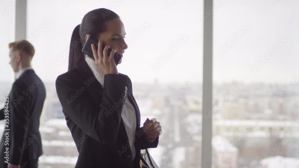 Slowmo medium shot of young businesswoman standing in office building with panoramic window and making phone call. Businessmen in suits walking past her Slowmo medium shot of young businesswoman standing in office building with panoramic window and making phone call. Businessmen in suits walking past her