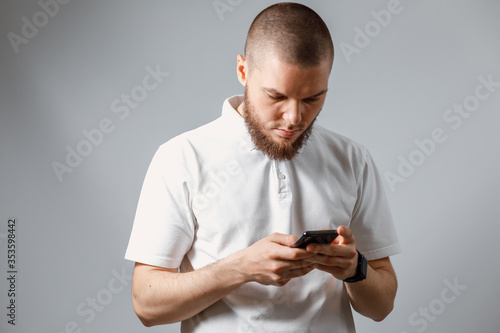 Portrait of a happy young man in a white T-shirt carefully looking at the phone on gray background. isolated, Copyspace © Yura