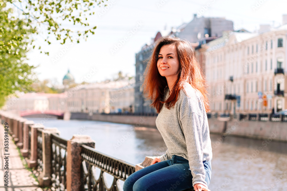 A brunette girl is sitting on the embankment on a Sunny day, green trees everywhere, walking around the city