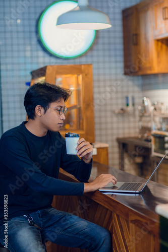 Young asian man drinking coffee in cafe and using laptop computer. Freelancer working from coffee shop