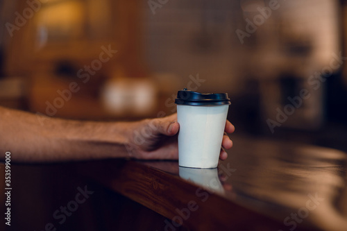 Takeaway paper cup of coffee on wooden table in cafe. Close up of man's hand holding coffee to go in coffee shop