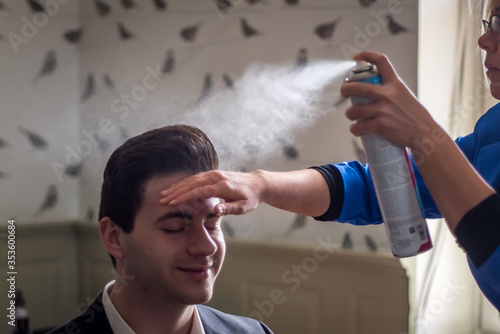A colourful photo of a busy professional hairdresser, she is preparing the groom ready for the big day wedding ceremony. The hairdressor is holding a hair spray can in her hand.