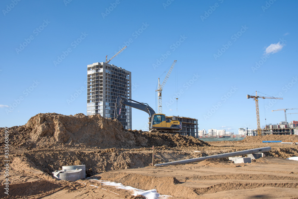 Tower cranes constructing residential buildings. Excavator on earthmoving at construction site. Backhoe dig ground for foundation and laying sewer pipes district heating. Tower cranes constructing residential buildings. Excavator on earthmoving at construction site. Backhoe dig ground for foundation and laying sewer pipes district heating.