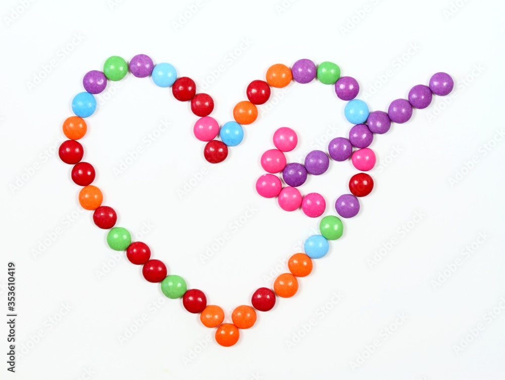 Sweet heart made from colored smarties with Cupido arrow. Chocolate candies in a shape of heart on white background, top view. Sweet heart made from colored smarties with Cupido arrow. Chocolate candies in a shape of heart on white background, top view.