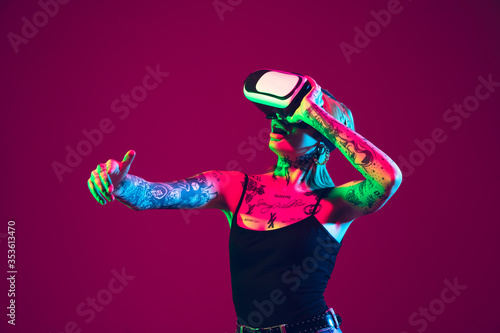Playing with VR-headset. Young caucasian woman on purple studio background in neon light. Beautiful female model with tattoos. Human emotions, facial expression, sales, ad concept. Freak's culture. © master1305