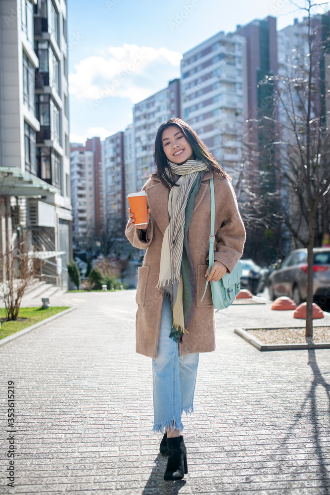 Fototapeta premium Brunette female in beige coat walking along the street with coffee cup, smiling