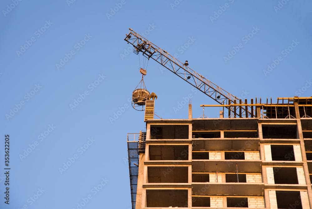Naklejka premium Tower cranes working at construction site against blue sky. Crane lifting a concrete bucket. Construction process of the new residential buildings. Transportation blocks and pouring of the cement mix