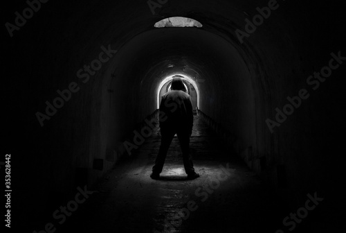 The figure of a man in a hood in a dark gloomy underground tunnel  standing in front of the light and lit through a hole in the ceiling.