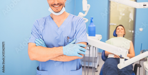 Young smiling handsome male doctor in blue medical uniform, disposable medical facial mask with equipment in dentistry office and happy patient in the dental chair. Stomatology concept. Copy space