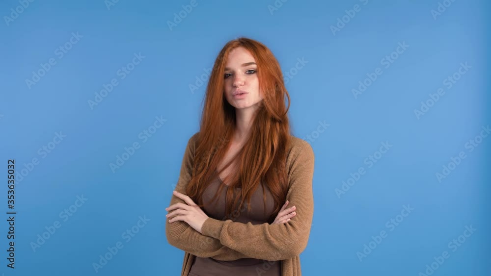Ginger lady in brown t-shirt and cardigan. She is flirting, winking, smiling and kissing you while posing against blue studio background. Close up