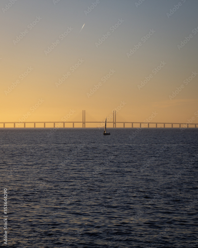 A single sailing boat is out on the the calm ocean between Denmark and Sweden during sunset with the Oresund bridge in the background