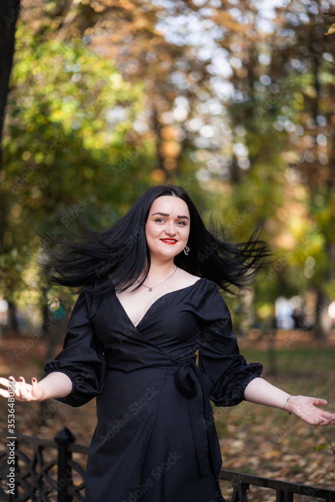 Autumn fashion outdoor. The girl with red lips in fashionable stylish black dress, autumnal lifestyle on the background of blurry yellow-green trees in the park. Vertical Autumn fashion outdoor. The girl with red lips in fashionable stylish black dress, autumnal lifestyle on the background of blurry yellow-green trees in the park. Vertical