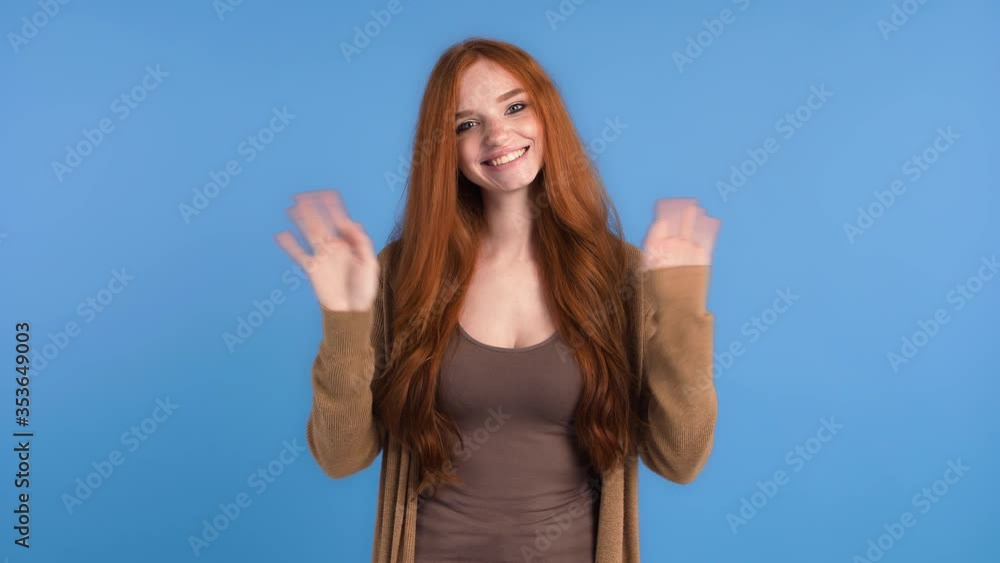 Redhead girl in brown t-shirt and cardigan. She is waving her hands, saying hi and smiling while posing against blue studio background. Close up Redhead girl in brown t-shirt and cardigan. She is waving her hands, saying hi and smiling while posing against blue studio background. Close up