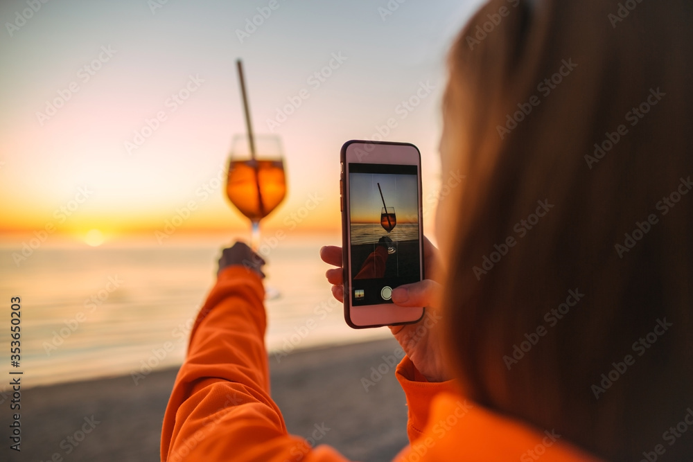 Fototapeta premium Girl Holding Glass of Drink on Beach at Beautyfull Romantic Sunset Taking Pictures with Phone. Glass and Hand Close Up. View From Back