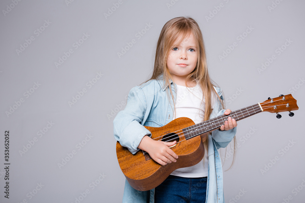 Fototapeta premium baby girl playing music on a small guitar. European girl with a guitar on a gray background. The concept of leisure and creativity
