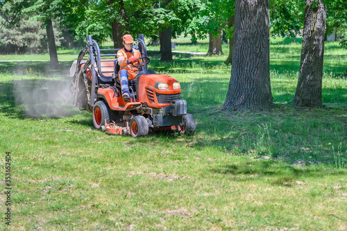 Man mows the grass under trees in park by riding mowing machine in sunny day