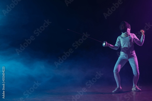 Swordswoman in fencing suit and mask holding rapier on black background with smoke and lighting