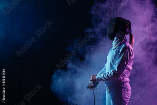 Side view of fencer in fencing mask holding rapier on black background with smoke