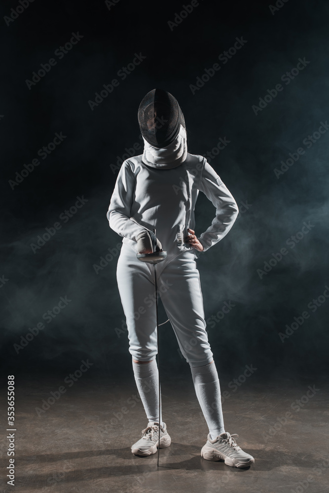 Fencer with hand on hip holding rapier on black background with smoke
