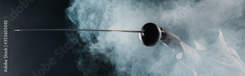 Fotografía Panoramic shot of fencer holding rapier on black background with smoke