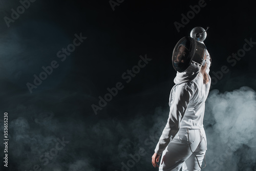 Back view of swordswoman in fencing mask looking at camera on black background with smoke