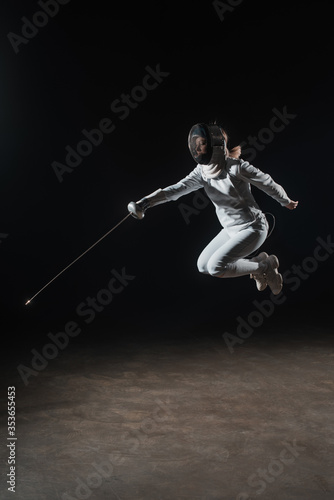 Fencer in fencing mask holding rapier and jumping on black background