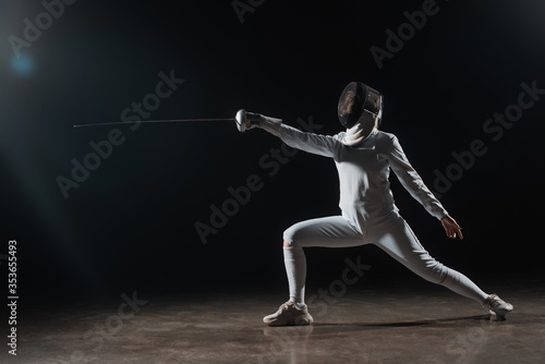 Fencer in fencing mask doing lunge while training with rapier on black background