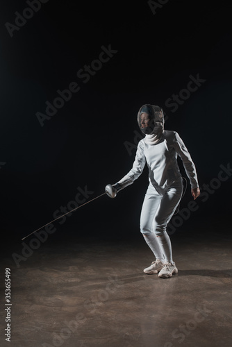 Swordswoman in fencing suit and mask training with rapier on black background