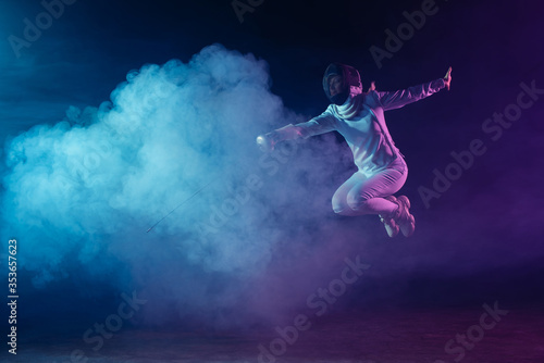 Fencer jumping while exercising with rapier on black background with lighting and smoke