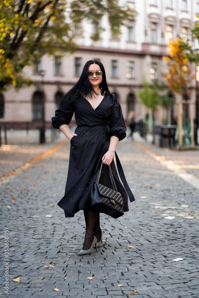 Autumn fashion outdoor. The girl with red lips in fashionable stylish black dress and sunglasses, autumnal lifestyle on the background of blurry yellow-green trees in the park. Vertical