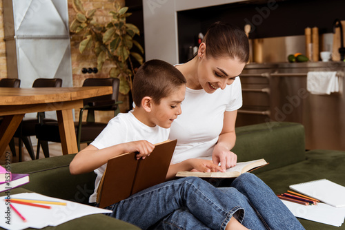 Selective focus of boy and smiling mother pointing with fingers while reading books near color pencils and paper at home