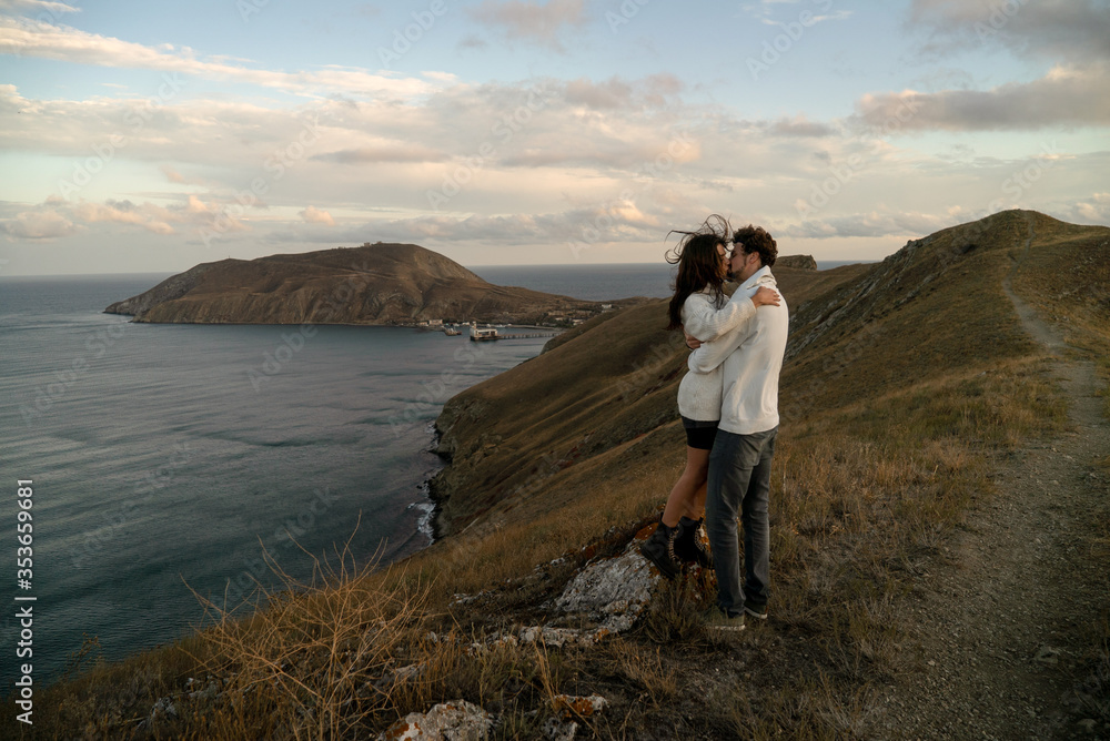 Sensual couple in white hoodies hugs and passionately kisses each other on the hill with a view to picturesque bay, beautiful clouds in a fall golden hour.