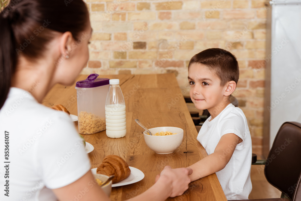 Selective focus of boy and mother holding hands near cereals and croissants on kitchen table Selective focus of boy and mother holding hands near cereals and croissants on kitchen table