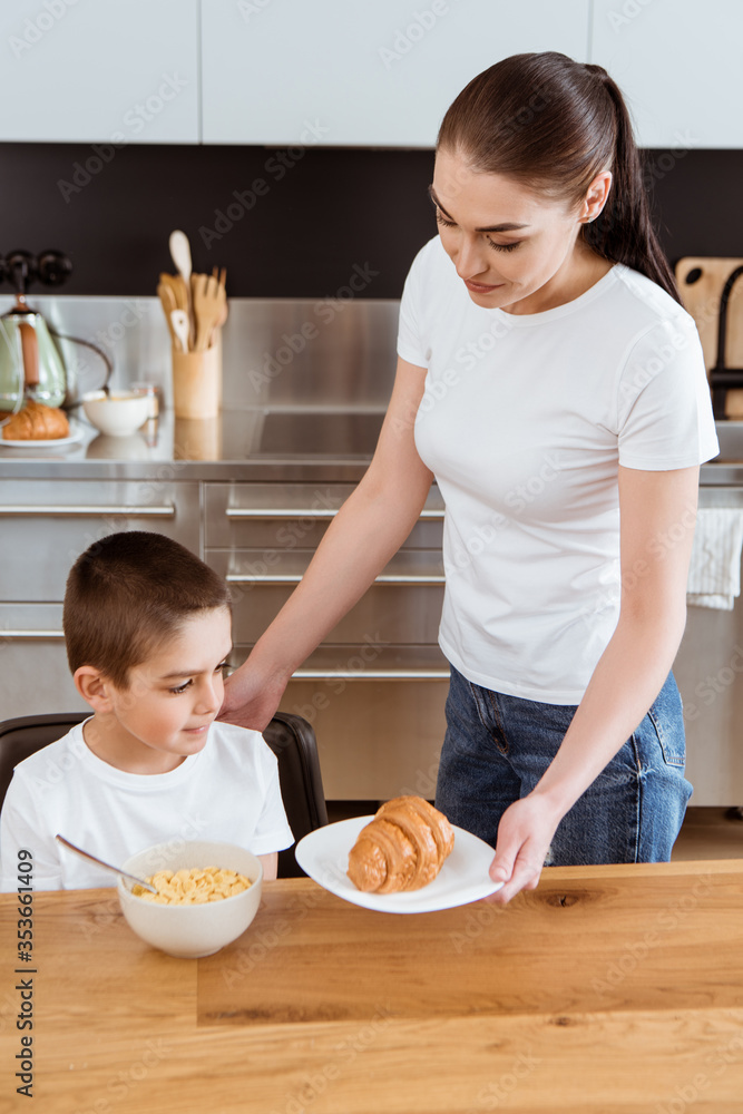Mother holding croissant near son and bowl of cereals on kitchen table Mother holding croissant near son and bowl of cereals on kitchen table