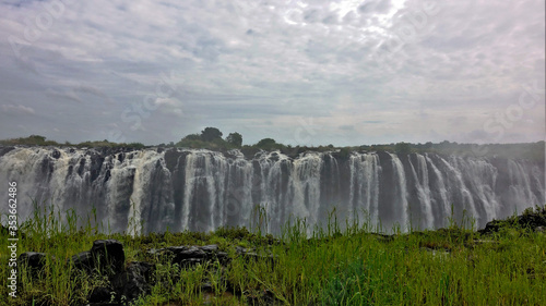 Unique Victoria Falls. The Zambezi River brings down numerous powerful streams of water through the edge of the gorge. In the foreground is another edge, covered with bright green grass. Cloudy.