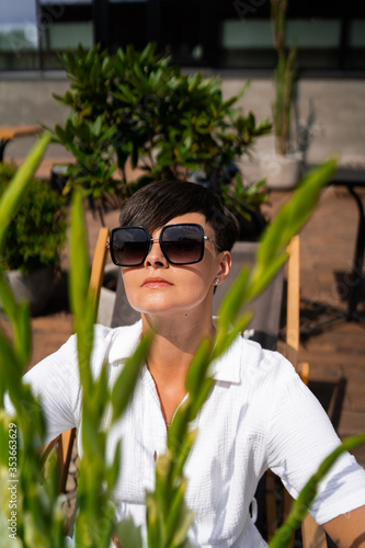 Beautiful woman with short brunette hair in white clothes and sunglasses. Fashion photography. Fashion model posing with flowers on wood background.