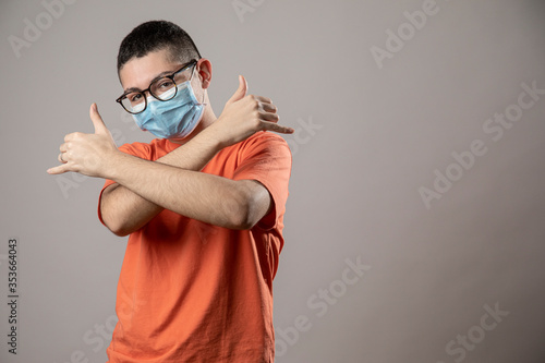 Ragazzo giovane con mascherina protettiva facciale, occhiali da vista neri e shirt arancione fa dei gesti da repper , isolato su sfondo grigio photo