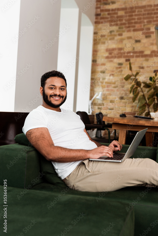 cheerful african american freelancer sitting on sofa with laptop and looking at camera - obrazy, fototapety, plakaty 