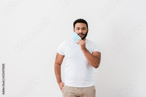 bearded african american man touching blue medical mask and standing with hand in pocket near white wall