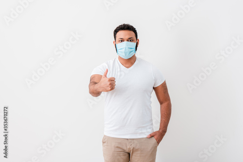 african american man in blue medical mask standing with hand in pocket and showing thumb up near white wall