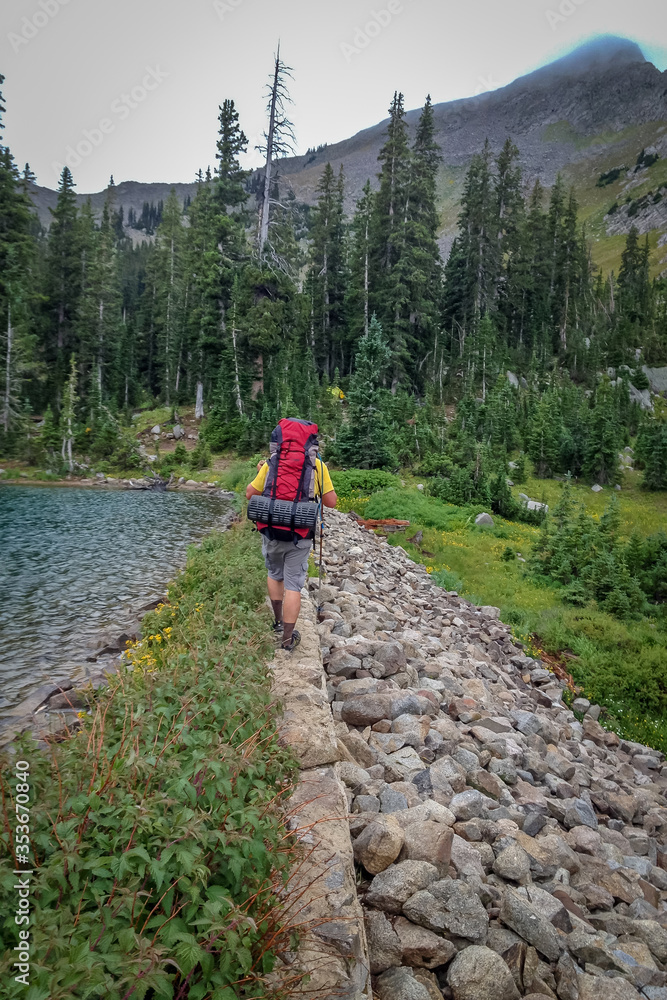 A man hiking in the mountains on a path next to a lake wearing a red backpack