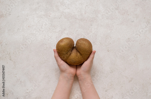 Ugly kiwi in the heart shape on a 
light background 
in children's hands. Funny, unnormal fruit or food waste concept. Horizontal orientation. Copy space. photo