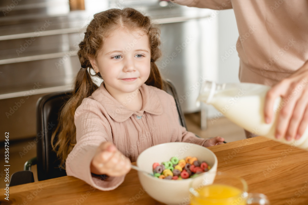 selective focus of mother holding bottle with milk near bowl with tasty corn flakes and cute daughter selective focus of mother holding bottle with milk near bowl with tasty corn flakes and cute daughter